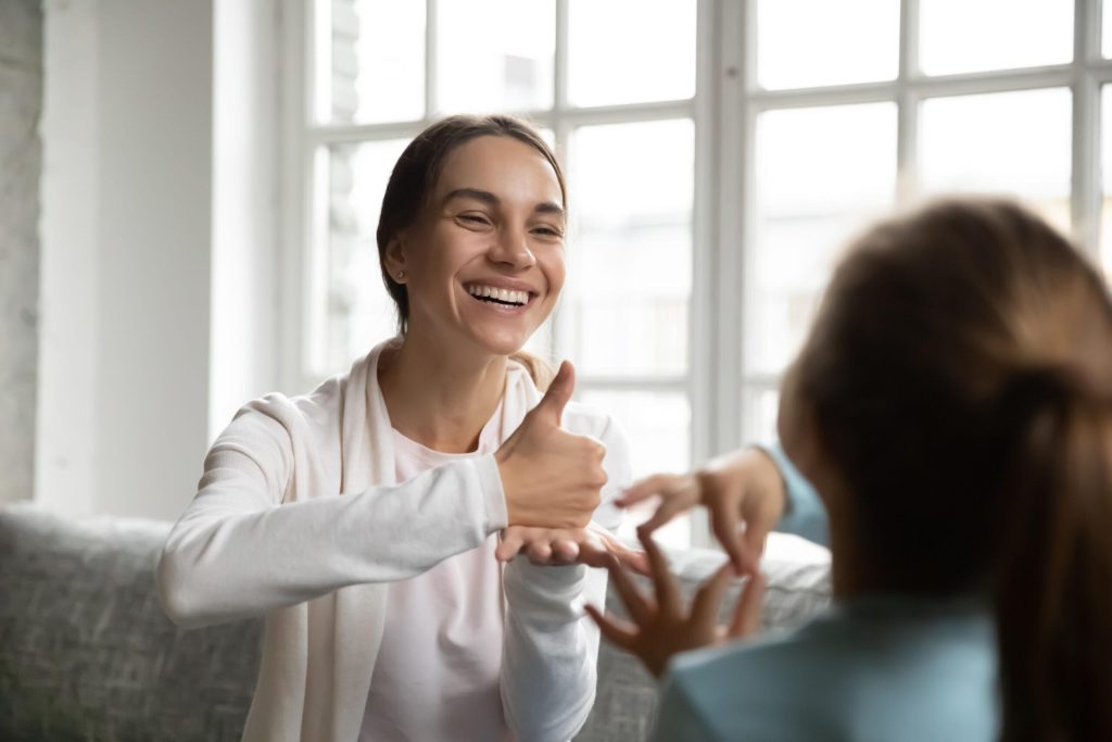 Smiling young Caucasian woman make hand gesture practice / nonverbal talk / sign language with child.